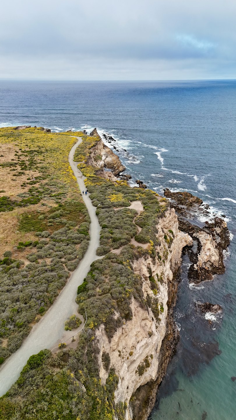 Two people walk along a winding coastal cliff path.