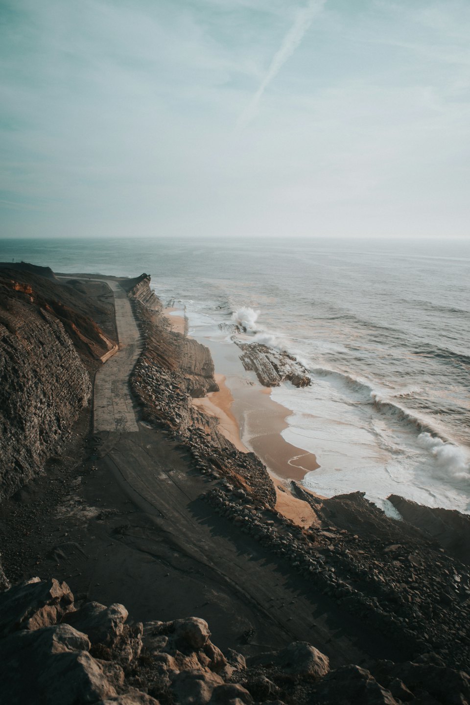 A winding coastal road follows cliffs above a sandy beach.