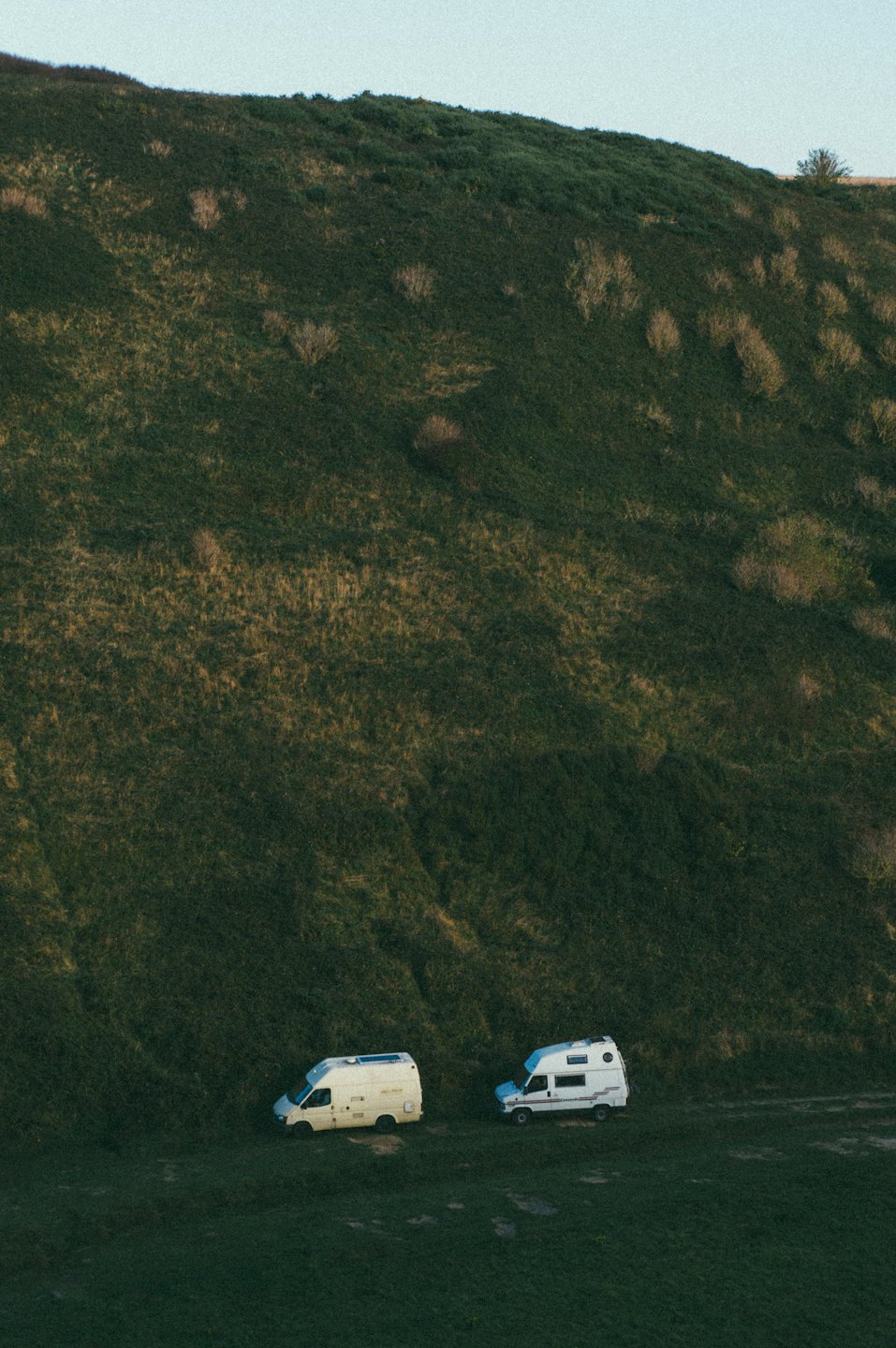 Two camper vans parked on grass at the base of a hillside.